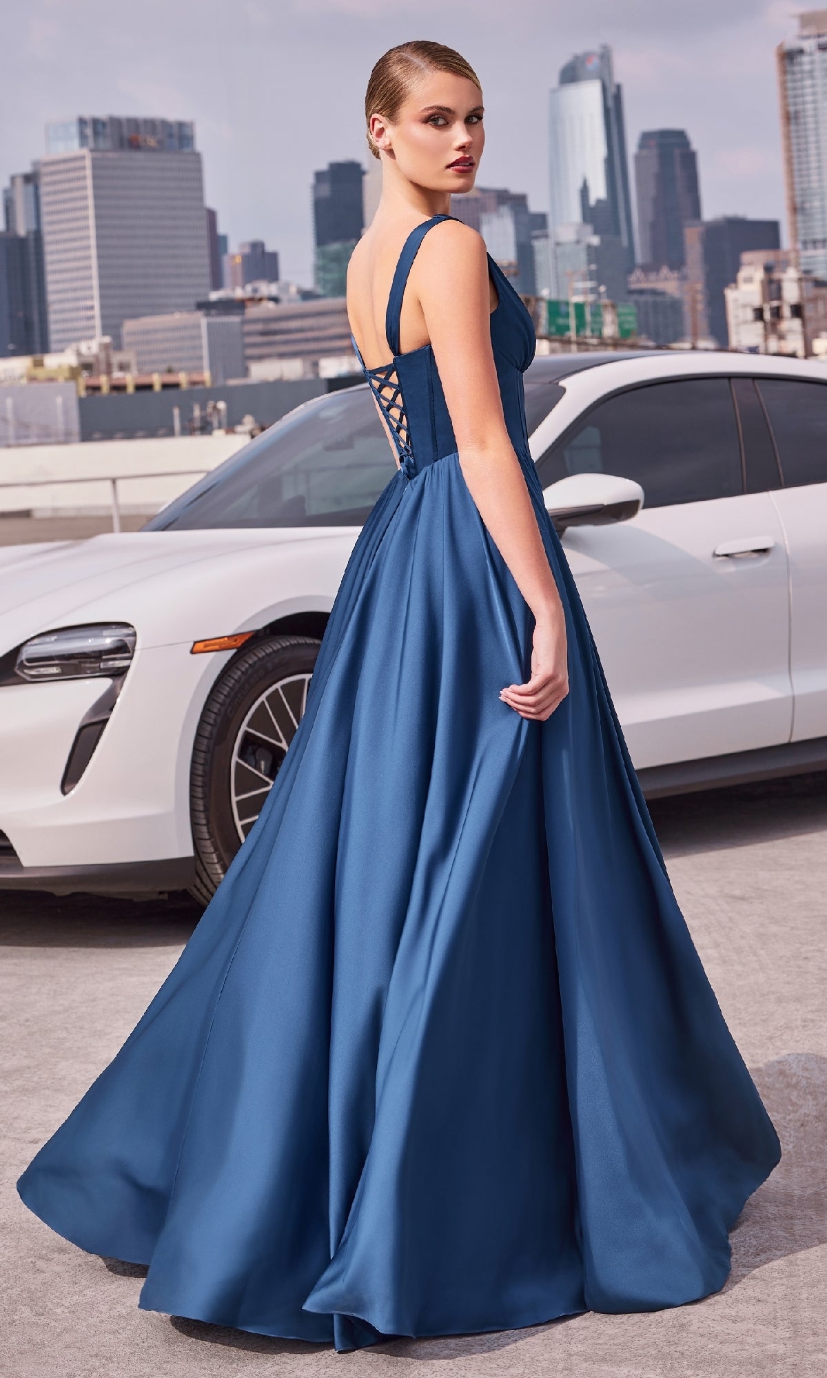 A woman wearing the Lace-Up Long A-Line Prom Dress Ladivine 7447 poses in front of a white car with a city skyline in the background.